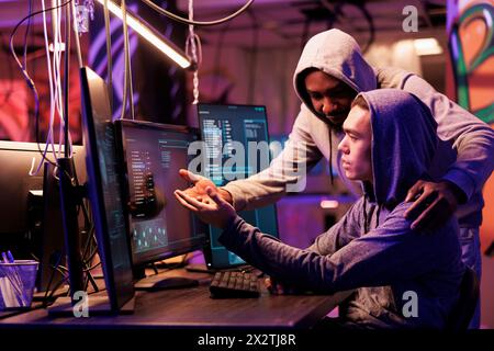 Two hackers collaborating while breaching data and coding malicious software on computer. Diverse criminals working together while programming ransomware in abandoned warehouse Stock Photo