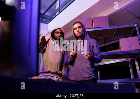 Hackers looking at computer and planning phishing attack. Criminals pointing at screen and discussing ransomware for fraud illegal activity in abandoned warehouse at night Stock Photo