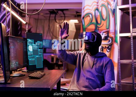 Hacker wearing vr glasses and getting unauthorized illegal access while exploring metaverse. African american criminal using virtual reality helmet to navigate database while breaking law Stock Photo