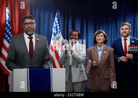 Successful mature male politician in formalwear standing by platform against group of intercultural delegates clapping their hands Stock Photo