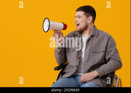 Young man in wheelchair shouting into megaphone on yellow background Stock Photo