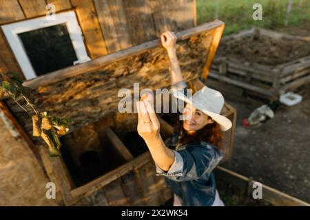 Happy middle-aged woman in hat collects fresh egg from a chicken coop at dawn. Stock Photo