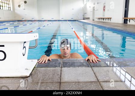 Caucasian young male swimmer wearing cap and goggles, resting at swimming pool edge Stock Photo