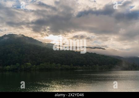 Morning sun breaks through heavy clouds above a forested ridge with mist at its base, reflected in a calm river near Glen Ferris, West Virginia. Stock Photo