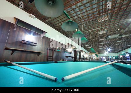 Two cues are on billiard table with green cloth inside club with metal lamps. Stock Photo