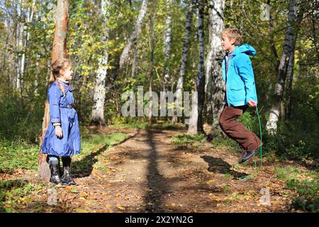 Happy boy jumps with skipping rope and girl looks at him in forest on sunny day. Stock Photo