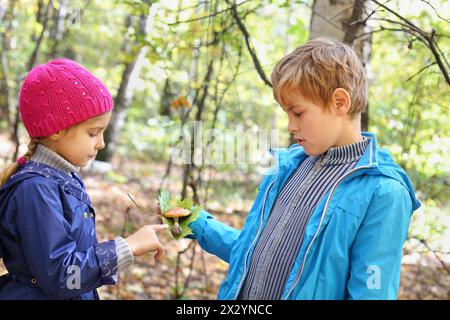 Boy in blue jacket holds green leaf and toadstool and shows it to girl in autumn forest. Stock Photo