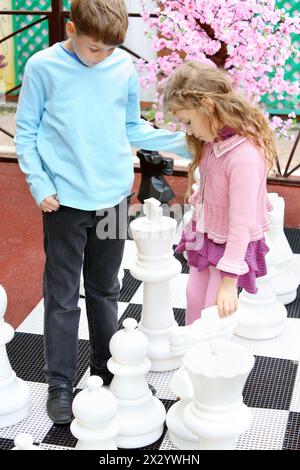 Boy and girl move big chess pieces on big chessboard in park. Stock Photo
