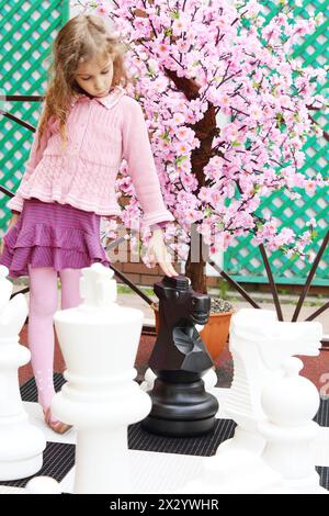 Girl next to artificial cherry blossom touches big chess pieces on big chessboard in park. Focus on tree. Stock Photo