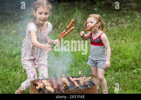 Two little girl making barbecue on the grill on nature, one girl eating a sausage on a skewer. Stock Photo