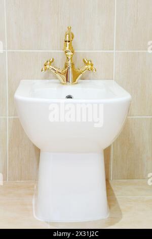 Clean and white bidet with gold-plated faucet in light bathroom. Stock Photo