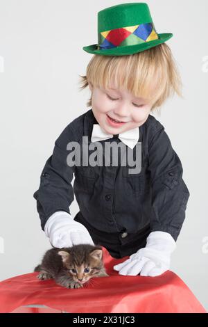 A little boy in a green hat and white gloves stroking a kitten on the table Stock Photo