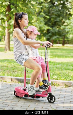 Teenage girl rides scooter, holding little girl on lap Stock Photo