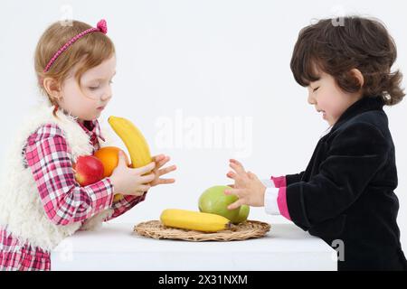 Little boy and girl stand and take fruits from white table on white background. Stock Photo