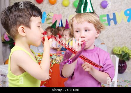 Two boys blow in party blowers at birthday party and three girls fun. Inscription Happy Birthday on wall. Focus on boys. Stock Photo