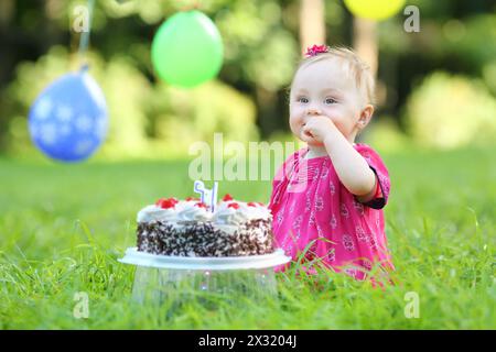 Little girl in red dress eating cake on her first birthday in the park Stock Photo