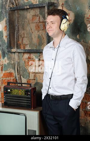 Smiling man in big headphones listens old radio in very old house. Stock Photo