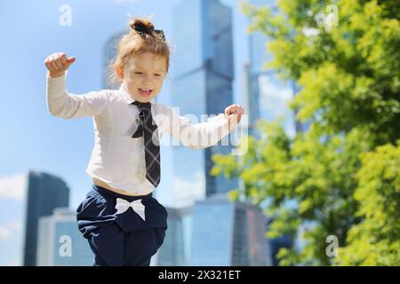 Little smiling girl  stands near tree near skyskrapers at sunny day. Stock Photo