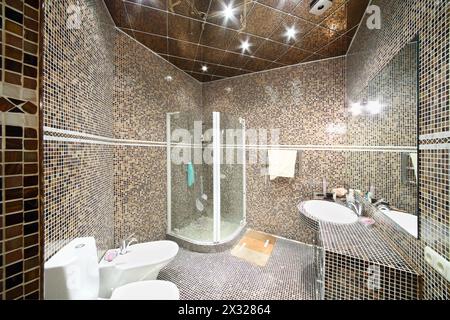 Small bathroom with shower unit, toilet and sink with small black and brown tiles. Stock Photo