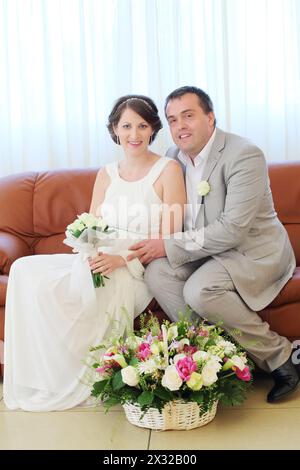 Beautiful bride and groom sit on the couch with a basket of flowers at the feet Stock Photo