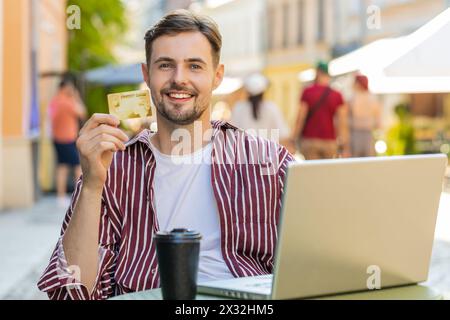 Man using credit bank card laptop computer while transferring money, purchases online shopping, order food delivery booking hotel room outdoors. Young guy tourist sitting at table in city cafeteria Stock Photo