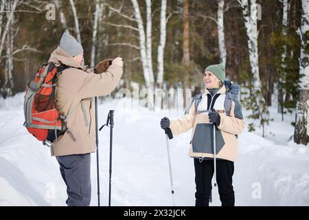 Medium long shot of senior Caucasian tourist holding smartphone taking photo of his wife in forest park on winter day Stock Photo