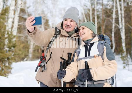 Medium shot of cheerful senior couple in sportswear with backpacks taking selfie photos while hiking in forest on winter day Stock Photo