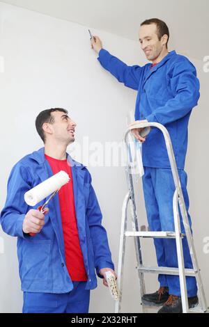 Happy workers in blue with construction tools, stand on ladder and look at each other in new apartment Stock Photo