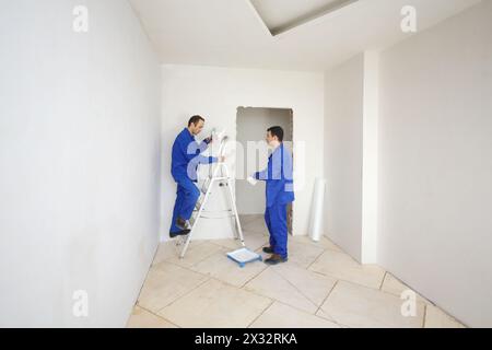 Two workers in blue clothes paint walls on ladder in new apartment Stock Photo