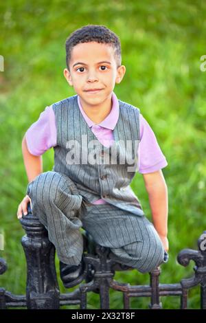 Little boy dressed in suit with vest sits on black wrought-iron fence Stock Photo