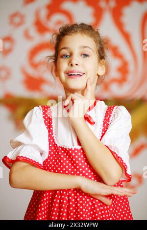 Beautiful girl in red folk costume poses and smiles near wall with pattern Stock Photo