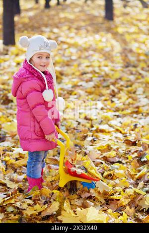 Little girl carries plastic wheelbarrow loaded with dried leaves in autumn park Stock Photo