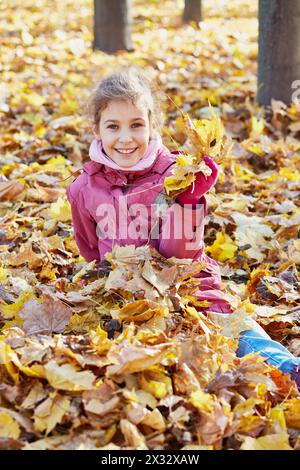 Smiling little girl in red jacket sits in drift of maple fallen yellow leaves in autumn park Stock Photo