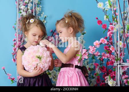 Two cute little girls in dresses make up bouquet of roses in room with flowers. Stock Photo