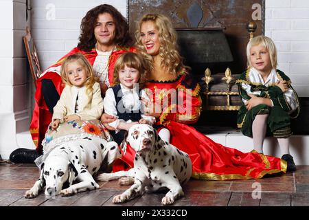 Smiling father, mother, little daughter and two sons in medieval costumes sit with two dalmatians near chimney with boiler. Stock Photo