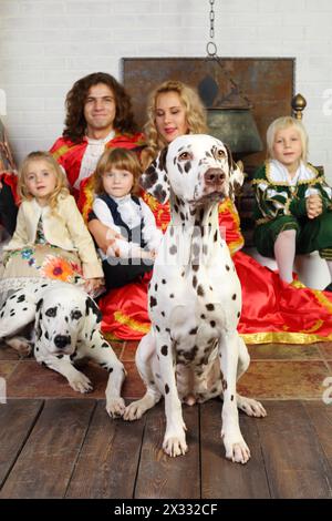 Father, mother, little daughter and two sons in medieval costumes sit with two dalmatians near chimney with boiler. Focus on right dog. Stock Photo
