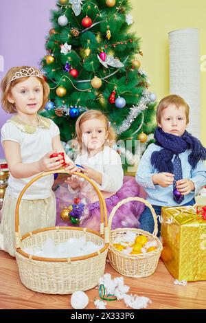 Two little girls and one boy sit under decorated christmas tree holding decorations in their hands Stock Photo