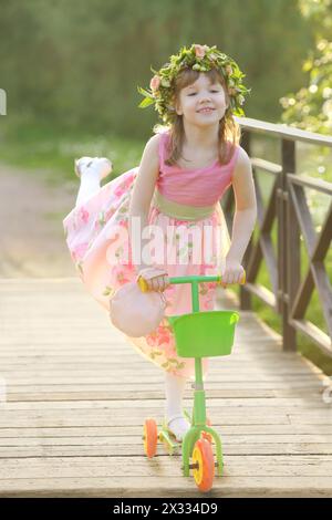 Little pretty girl in pink rides scooter on bridge in summer park. Stock Photo