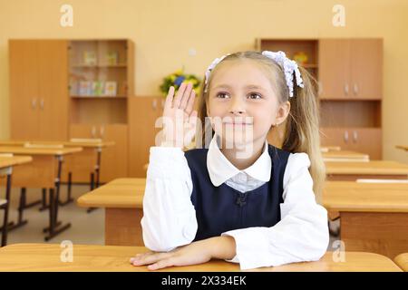 Little girl in school uniform sits at wooden school desk and raises hand to answer in classroom at school. Stock Photo