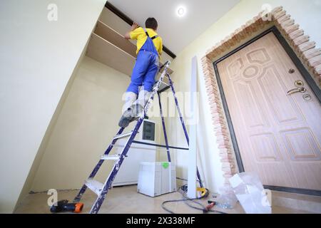 Man standing on ladder fixes shelves to wall for closet in hallway, bottom view Stock Photo