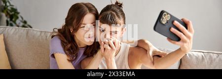 Two women in comfy attire sit on a couch, engrossed in a cell phone. Stock Photo