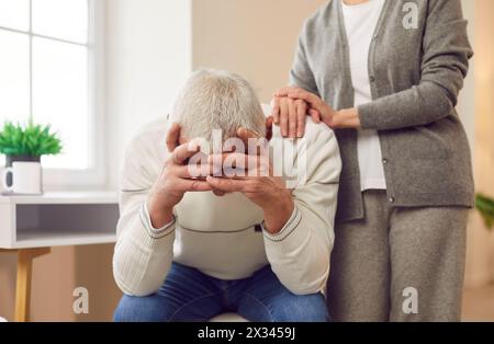 Senior woman trying to make peace with her upset elderly husband after quarrel sitting at home. Stock Photo