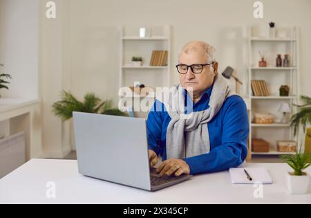 Old gray-haired senior mature man working using laptop sitting at the desk at home. Stock Photo
