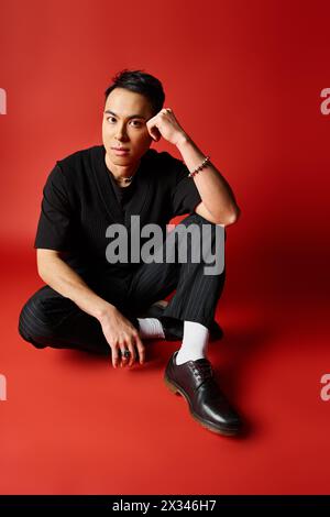 Handsome Asian man in black attire sitting on the ground, wearing black shoes, against a bold red studio background. Stock Photo