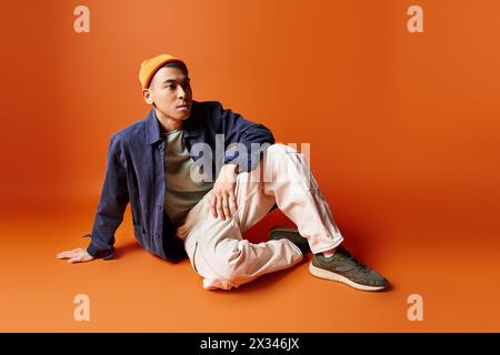 A handsome Asian man in stylish attire sits on the ground wearing a hat, against an orange background in a studio setting. Stock Photo