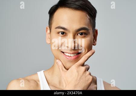 A handsome Asian man with a smile on his face, exuding happiness and confidence in a grey studio while using skincare products. Stock Photo