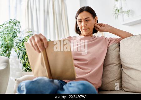 A mature woman in cozy homewear sitting on a couch, engrossed in a book. Stock Photo