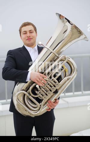 Man in black suit poses with tuba on roof of tall building at winter day Stock Photo