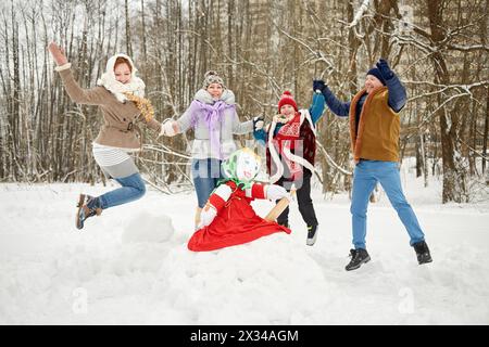 Happy family of four jumps holding hands around stuffed dummy Maslenitsa in winter park. Stock Photo