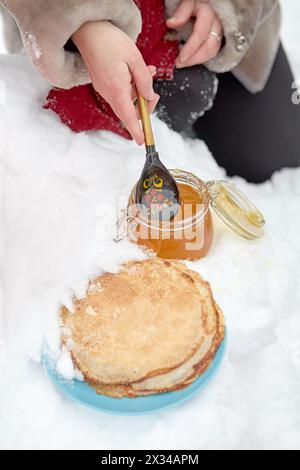 Female hand takes honey with wooden spoon from glass jar and pours it onto pancakes in plate on snow. Stock Photo